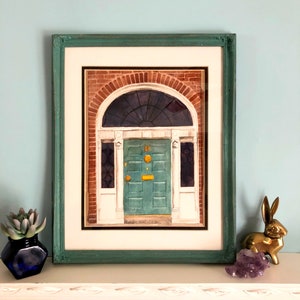 May include: A framed watercolour painting of a green door with a gold door knocker. The door is set in a brick building with a white archway above it.