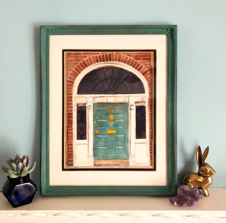 May include: A framed watercolour painting of a green door with a gold door knocker. The door is set in a brick building with a white archway above it.