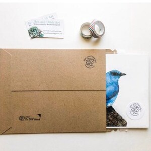 May include: A brown paper envelope containing a white card with a blue bird illustration. The card features a small round sticker with a floral design. The envelope has a return address label with the text "Bloom and Chick Art".