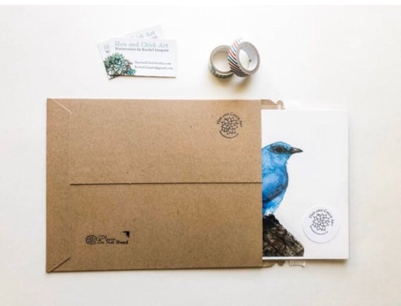 May include: A brown paper envelope containing a white card with a blue bird illustration. The card features a small round sticker with a floral design. The envelope has a return address label with the text "Bloom and Chick Art".