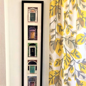 May include: A black framed picture with five photos of different doors. Each photo is in a white mount. The doors are all different colours and styles. The picture is hanging on a white wall next to a yellow and grey patterned curtain.