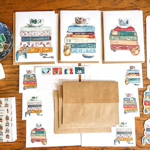 May include: A stationery set featuring greeting cards, envelopes, and stickers with watercolor illustrations of stacked books topped with teacups. The collection includes an address book, a bookmark, and loose sheets of paper with book-themed motifs.