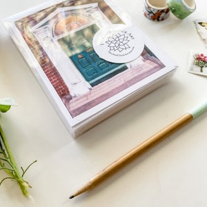 May include: A stack of blank greeting cards with a watercolor illustration of a teal door with a stained glass window. The cards are wrapped in clear plastic and have a white circle sticker with the text "Hen and Chick Art" and a floral design.