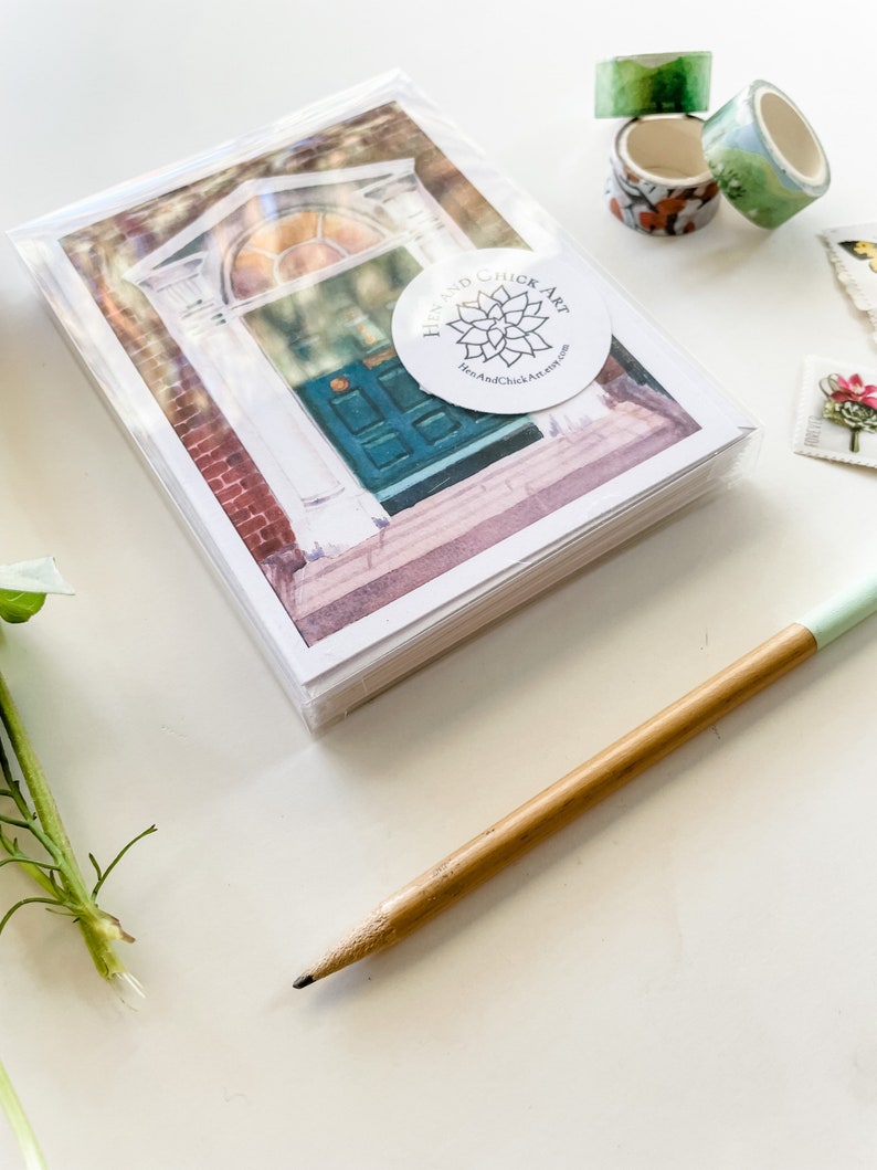 May include: A stack of blank greeting cards with a watercolor illustration of a teal door with a stained glass window. The cards are wrapped in clear plastic and have a white circle sticker with the text "Hen and Chick Art" and a floral design.