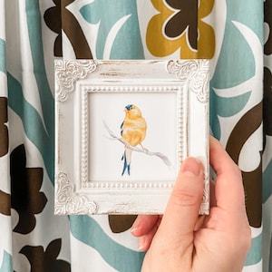 Pode incluir: Uma pequena pintura em aquarela com moldura branca, retratando um pássaro amarelo empoleirado em um galho. O pássaro tem cabeça e penas da cauda azuis. A moldura tem detalhes ornamentados. O fundo é uma cortina estampada.