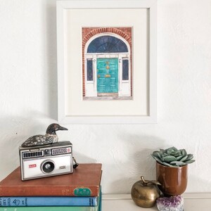 May include: A framed watercolour painting of a teal green door with a brass door knocker and a small window above the door. The door is set in a brick building with white trim. The painting is on a white background.