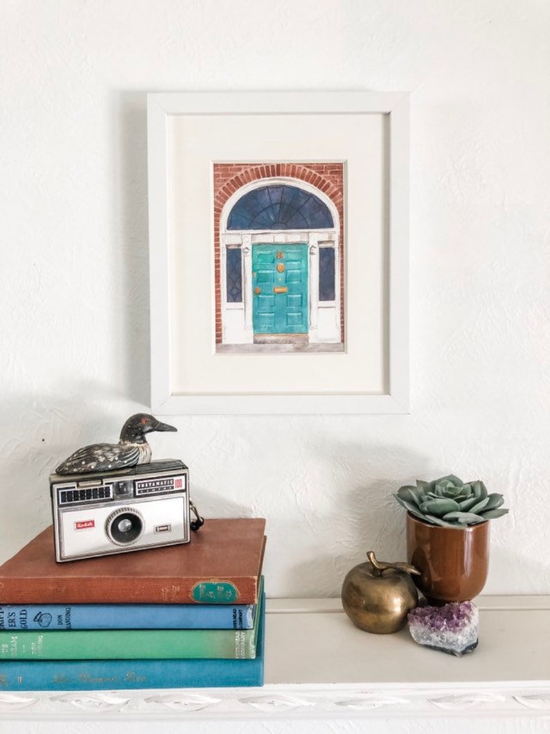 May include: A framed watercolour painting of a teal green door with a brass door knocker and a small window above the door. The door is set in a brick building with white trim. The painting is on a white background.