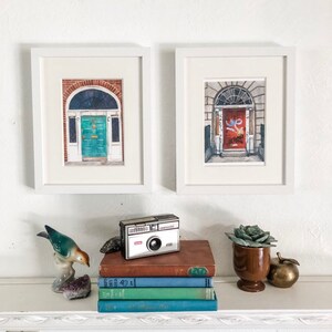 May include: Two framed watercolour paintings of doors in Dublin, Ireland. The paintings are in white frames and are hanging on a white wall. A vintage camera, a stack of books, a succulent plant in a brown pot, and a gold apple are on a shelf below the paintings.