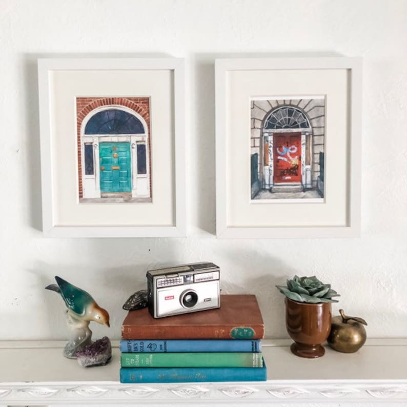 May include: Two framed watercolour paintings of doors in Dublin, Ireland. The paintings are in white frames and are hanging on a white wall. A vintage camera, a stack of books, a succulent plant in a brown pot, and a gold apple are on a shelf below the paintings.