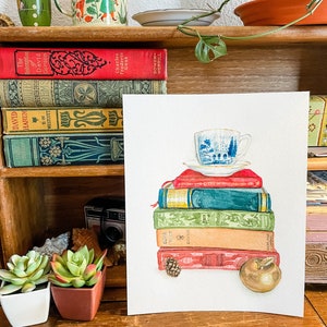 May include: Watercolour painting of a stack of vintage books with a teacup and saucer on top. The books are various colours and have titles such as "David Harum" and "The Redemption of David Corson". There is a small pine cone and a red apple at the bottom of the stack.