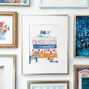 May include: A watercolour painting of a stack of books with a teacup and saucer on top. A blue and white Dala horse is in front of the books. There are two keys on the table below the books. The painting is framed in a silver frame.
