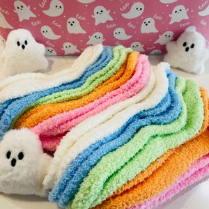 K&ouml;nnte beinhalten: Ein Stapel bunter, flauschiger Socken in Rosa, Orange, Gr&uuml;n, Blau und Wei&szlig;. Die Socken sind in einem Regenbogenmuster angeordnet. Wei&szlig;e Pl&uuml;sch-Geisterfiguren sind um die Socken platziert. Der Hintergrund ist rosa mit wei&szlig;en Geisterillustrationen und dem Wort "boo".