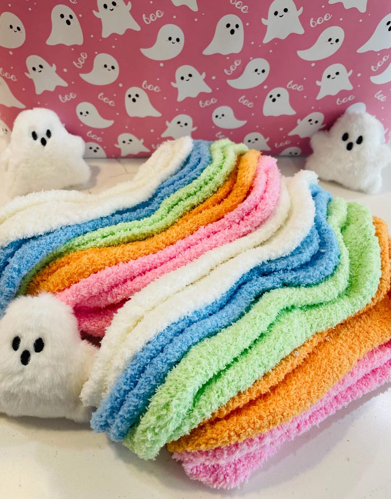 K&ouml;nnte beinhalten: Ein Stapel bunter, flauschiger Socken in Rosa, Orange, Gr&uuml;n, Blau und Wei&szlig;. Die Socken sind in einem Regenbogenmuster angeordnet. Wei&szlig;e Pl&uuml;sch-Geisterfiguren sind um die Socken platziert. Der Hintergrund ist rosa mit wei&szlig;en Geisterillustrationen und dem Wort "boo".