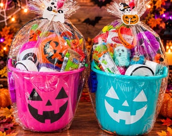 Könnte beinhalten: Zwei Halloween-Eimer aus Kunststoff, einer in leuchtendem Pink und einer in Türkis, jeweils mit einem weißen Kürbisgesicht. Die Eimer sind in durchsichtige Folie eingewickelt und mit kleinen Spielzeugen und Süßigkeiten gefüllt, verziert mit einem Geist- oder Kürbisanhänger.