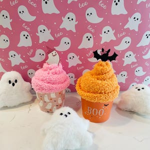 K&ouml;nnte beinhalten: Eine Halloween-Dekoration mit Pl&uuml;sch-Geisterfiguren und cupcake-f&ouml;rmigen Beh&auml;ltern. Ein Cupcake ist rosa, ein anderer orange mit dem Text "BOO". Der Hintergrund ist rosa mit Geisterillustrationen.