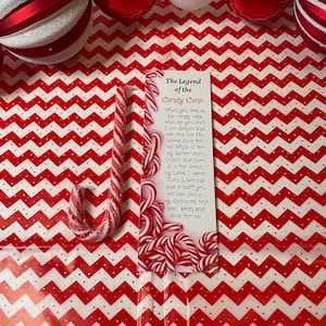 May include: A candy cane with red and white stripes, next to a bookmark with the text "The Legend of the Candy Cane." The background features a red and white zigzag pattern and Christmas ornaments.