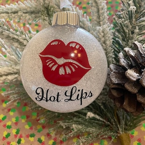 May include: A silver glitter Christmas ornament with a red lip print and the text "Hot Lips" in black.