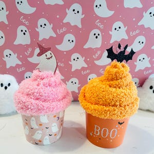 K&ouml;nnte beinhalten: Zwei dekorative Becher zum Thema Halloween. Einer ist rosa mit einem Geisterdesign und einem Hexenhut-Topper. Der andere ist orange mit dem Text "BOO" und einem Fledermaus-Topper. Der Hintergrund zeigt ein rosa Geistermuster.