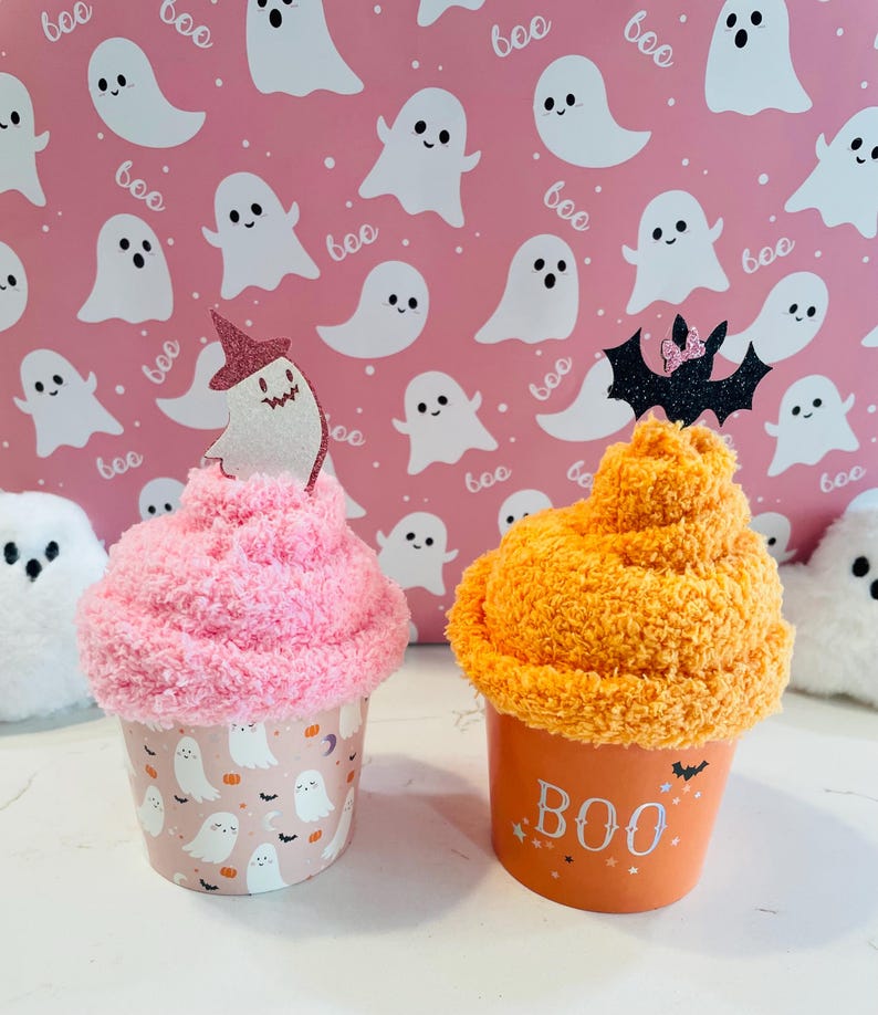 K&ouml;nnte beinhalten: Zwei dekorative Becher zum Thema Halloween. Einer ist rosa mit einem Geisterdesign und einem Hexenhut-Topper. Der andere ist orange mit dem Text "BOO" und einem Fledermaus-Topper. Der Hintergrund zeigt ein rosa Geistermuster.