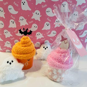 K&ouml;nnte beinhalten: Halloween-Geschenke mit Pl&uuml;sch-Geisterfiguren und cupcake-f&ouml;rmigen Artikeln. Ein Cupcake ist in Zellophan mit einem Etikett verpackt. Der Hintergrund ist rosa mit wei&szlig;en Geisterillustrationen und dem Wort "boo".