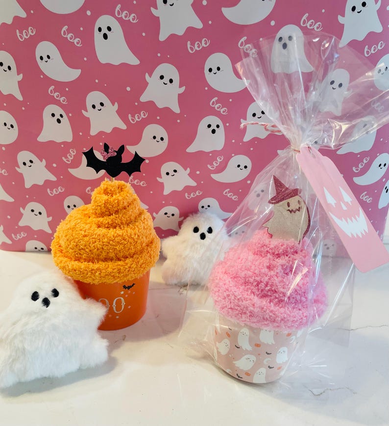 K&ouml;nnte beinhalten: Halloween-Geschenke mit Pl&uuml;sch-Geisterfiguren und cupcake-f&ouml;rmigen Artikeln. Ein Cupcake ist in Zellophan mit einem Etikett verpackt. Der Hintergrund ist rosa mit wei&szlig;en Geisterillustrationen und dem Wort "boo".