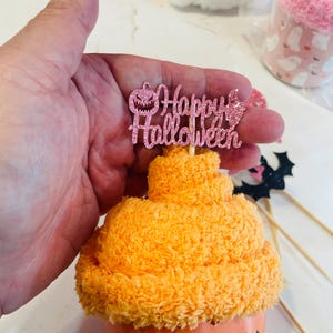 K&ouml;nnte beinhalten: Ein orangefarbener Halloween-Cupcake mit einem rosa Glitzer-"Happy Halloween"-Topper. Der Cupcake befindet sich in einer orangefarbenen Tasse mit einem "Boo"-Design. Weitere Halloween-Dekorationen sind im Hintergrund.