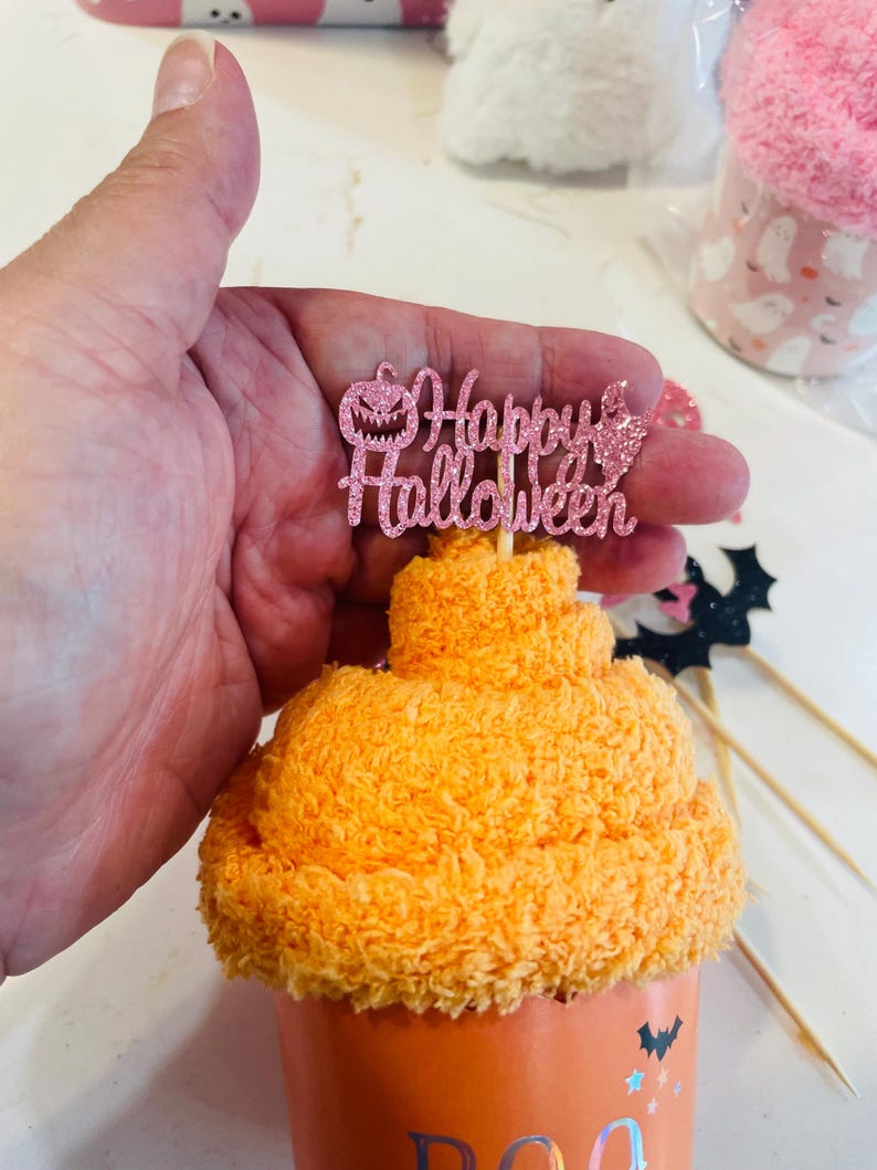 K&ouml;nnte beinhalten: Ein orangefarbener Halloween-Cupcake mit einem rosa Glitzer-"Happy Halloween"-Topper. Der Cupcake befindet sich in einer orangefarbenen Tasse mit einem "Boo"-Design. Weitere Halloween-Dekorationen sind im Hintergrund.