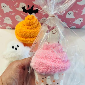 K&ouml;nnte beinhalten: Ein Halloween-Geschenkset mit einem rosa cupcake-f&ouml;rmigen Artikel mit Geisterdesign, verpackt in Zellophan. Weitere Artikel sind ein flauschiger orangefarbener Ball und ein wei&szlig;es Geisterpl&uuml;schtier, vor einem rosa Hintergrund mit Geistermustern.
