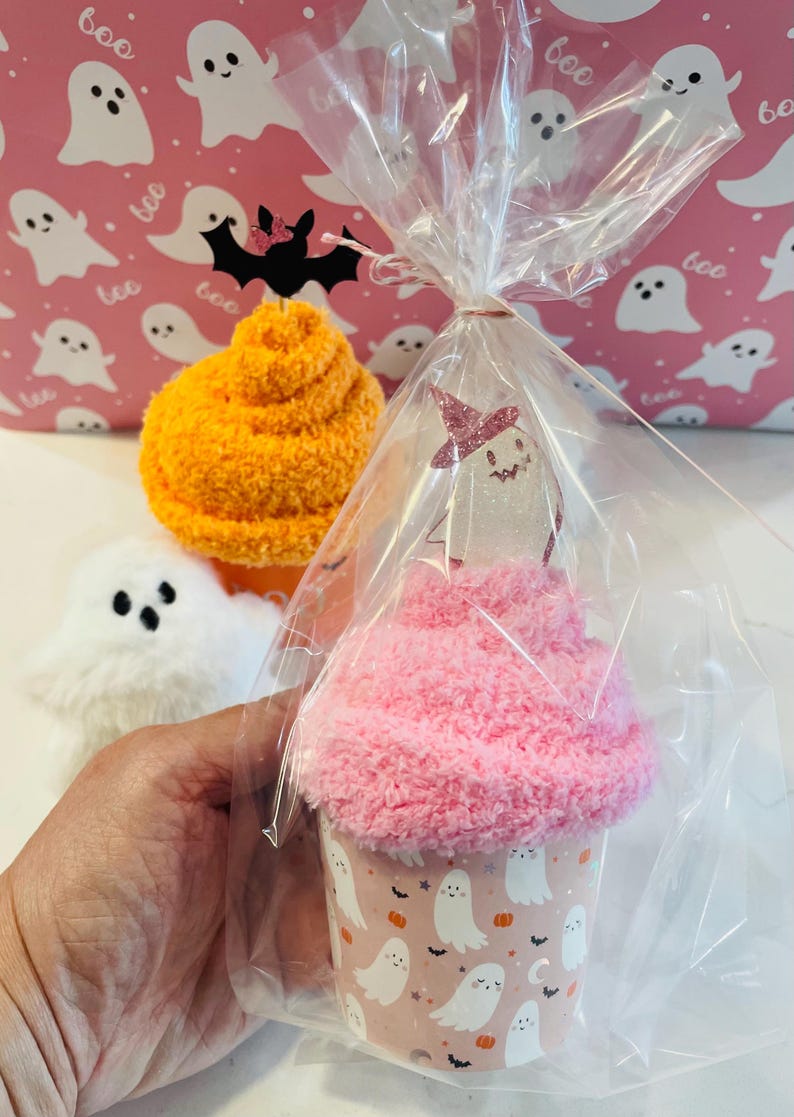 K&ouml;nnte beinhalten: Ein Halloween-Geschenkset mit einem rosa cupcake-f&ouml;rmigen Artikel mit Geisterdesign, verpackt in Zellophan. Weitere Artikel sind ein flauschiger orangefarbener Ball und ein wei&szlig;es Geisterpl&uuml;schtier, vor einem rosa Hintergrund mit Geistermustern.