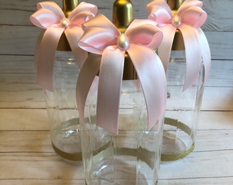 Könnte beinhalten: Drei durchsichtige Glasflaschen für Babys mit goldfarbenen Verschlüssen und Böden, die mit einem Band aus Goldglitter verziert sind. Jede Flasche ist mit einer hellrosa Satinschleife am Flaschenhals versehen, die eine weiße Perle trägt.