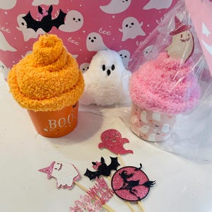 K&ouml;nnte beinhalten: Halloween-Geschenkset mit orangefarbenen und rosa cupcake-f&ouml;rmigen Artikeln, einem wei&szlig;en Pl&uuml;schgeist und glitzernden Toppern. Der orangefarbene Artikel tr&auml;gt das Wort "BOO". Der Hintergrund ist rosa mit wei&szlig;en Geisterillustrationen.