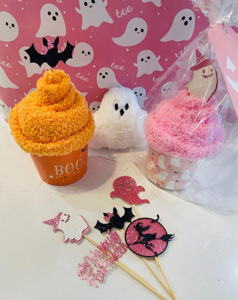 K&ouml;nnte beinhalten: Halloween-Geschenkset mit orangefarbenen und rosa cupcake-f&ouml;rmigen Artikeln, einem wei&szlig;en Pl&uuml;schgeist und glitzernden Toppern. Der orangefarbene Artikel tr&auml;gt das Wort "BOO". Der Hintergrund ist rosa mit wei&szlig;en Geisterillustrationen.