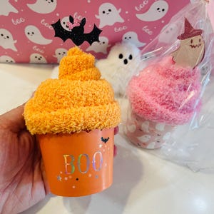 K&ouml;nnte beinhalten: Orangefarbener Halloween-Cupcake mit dem Wort "BOO" auf der Tasse. Der Cupcake ist mit einer schwarzen Glitzerfledermaus mit rosa Schleife gekr&ouml;nt. Ein rosa-wei&szlig;es Geschenk mit Geistermotiv im Hintergrund.