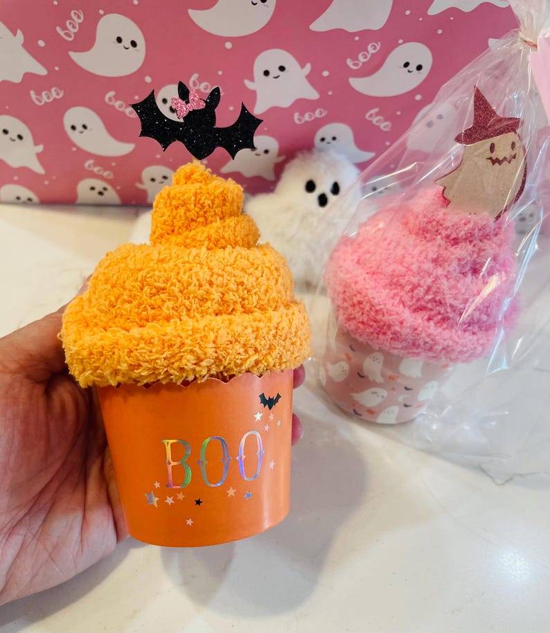K&ouml;nnte beinhalten: Orangefarbener Halloween-Cupcake mit dem Wort "BOO" auf der Tasse. Der Cupcake ist mit einer schwarzen Glitzerfledermaus mit rosa Schleife gekr&ouml;nt. Ein rosa-wei&szlig;es Geschenk mit Geistermotiv im Hintergrund.