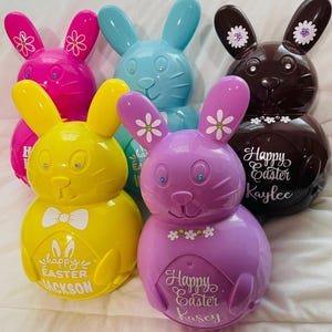 May include: Five Easter bunny shaped piggy banks in different colors. The bunnies are pink, blue, yellow, purple, and brown. Each bunny has a different name on it: Jackson, Kaylee, Kasey, and two others.