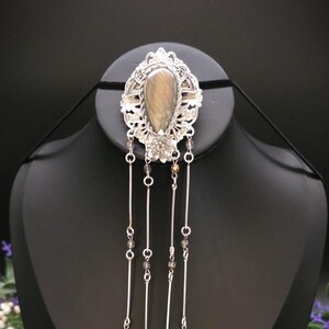 May include: A silver-toned brooch featuring an oval-shaped, brownish-grey stone set in an ornate filigree frame. Below the central piece, four dangling silver chains are adorned with small dark beads and finished with star-shaped charms.