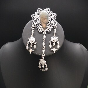 May include: A silver-toned brooch featuring a central teardrop-shaped grey stone set within an intricate filigree frame. Three dangling chains hang from the base, each adorned with small star-shaped charms and clear beads, creating a layered effect.