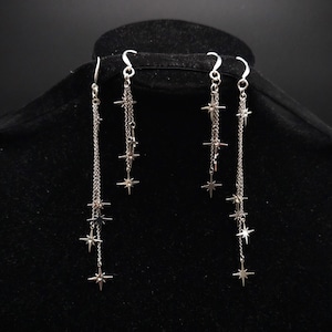 May include: Silver-toned dangle earrings featuring a series of star charms suspended from delicate chains. The earrings have a hook closure and are displayed on a black mannequin head. The stars are evenly spaced along the chains.