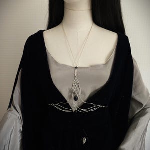 May include: A silver-toned necklace with an intricate, flowing design, featuring a dark, oval-shaped stone pendant. The necklace is displayed on a mannequin wearing a black velvet vest over a gray blouse, creating a contrast of textures and colors.