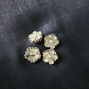 May include: Four silver-colored flower-shaped buttons with a glossy finish are arranged on a dark, textured surface. Each button features a detailed floral design with a central accent. The buttons are likely for sewing or crafting projects.