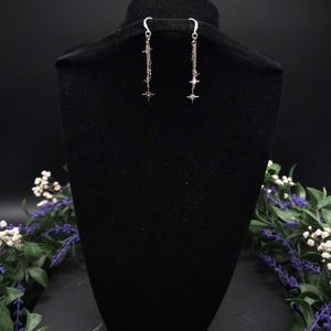 May include: Silver dangle earrings with a celestial theme. Each earring features a chain with multiple star charms and a larger star at the bottom. The earrings are displayed on a black velvet bust, with floral accents.