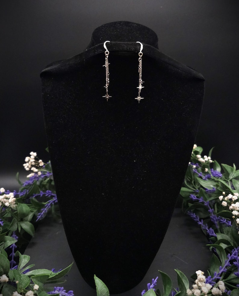 May include: Silver dangle earrings with a celestial theme. Each earring features a chain with multiple star charms and a larger star at the bottom. The earrings are displayed on a black velvet bust, with floral accents.