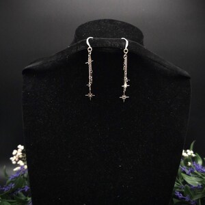 May include: Silver dangle earrings with starburst charms. The earrings hang from a curved hook and feature a chain design with small star accents. The earrings are displayed on a black velvet bust.