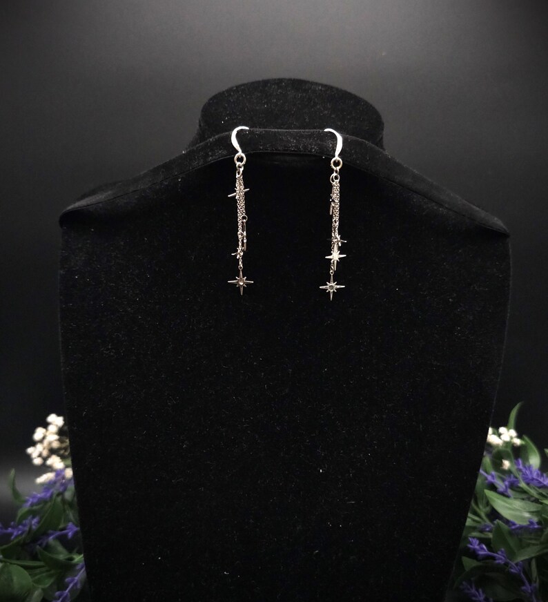 May include: Silver dangle earrings with starburst charms. The earrings hang from a curved hook and feature a chain design with small star accents. The earrings are displayed on a black velvet bust.