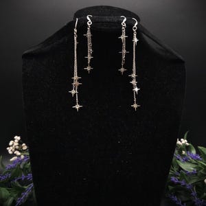 May include: Silver-toned dangle earrings featuring a chain design with multiple star charms. The earrings are displayed on a black mannequin head, showcasing their length and delicate design. The stars are evenly spaced along the chains.