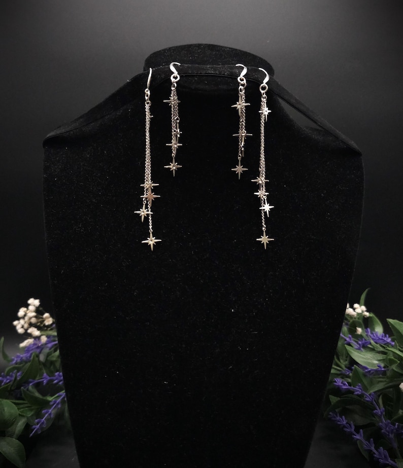 May include: Silver-toned dangle earrings featuring a chain design with multiple star charms. The earrings are displayed on a black mannequin head, showcasing their length and delicate design. The stars are evenly spaced along the chains.