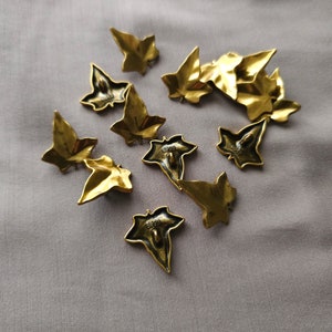 May include: A collection of ten gold-toned metal buttons shaped like ivy leaves. The buttons have a textured surface and are scattered on a light gray fabric background.