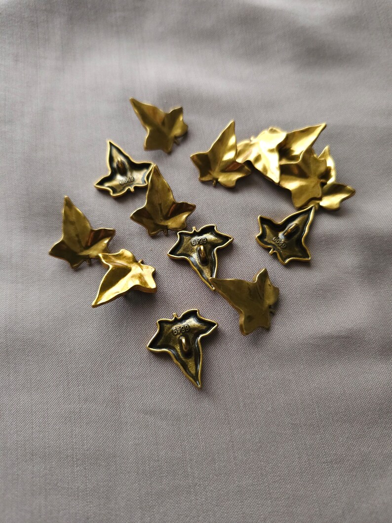 May include: A collection of ten gold-toned metal buttons shaped like ivy leaves. The buttons have a textured surface and are scattered on a light gray fabric background.