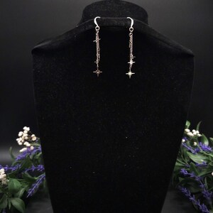May include: Silver dangle earrings with a star design. Each earring features a chain with multiple small stars and a larger star at the bottom. The earrings are displayed on a black velvet bust.
