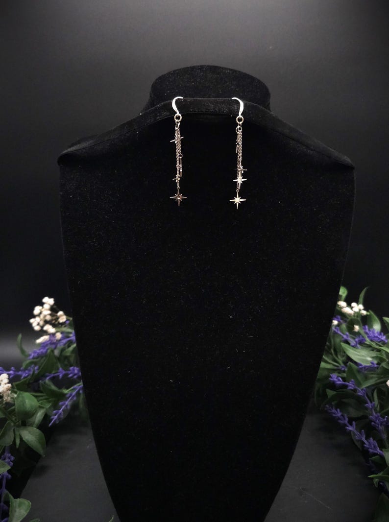 May include: Silver dangle earrings with a star design. Each earring features a chain with multiple small stars and a larger star at the bottom. The earrings are displayed on a black velvet bust.